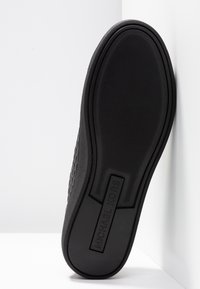 Semelle de chaussure noire Michael Kors avec grip texturé et nom de la marque moulé au centre, inclinée contre des murs blancs d’angle.