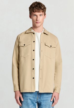 Chemise beige à boutons avec deux poches poitrine, boutons noirs et col. Portée sur un t-shirt blanc avec un jean en denim bleu clair.