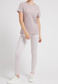 Světle růžové tričko s logem „Guess“, spárované s bílými legínami s jemným vzorem a bílými teniskami s šedými akcenty.