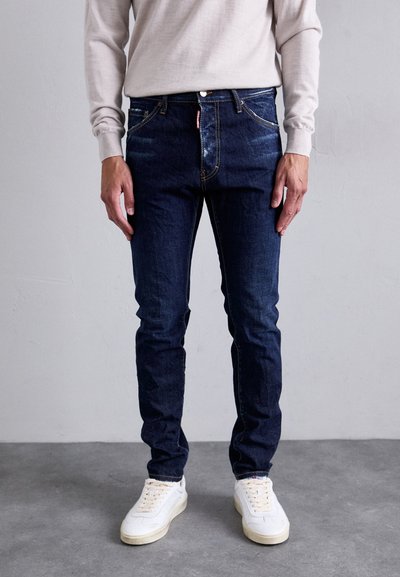 Person in dunkelblauen Slim-Fit-Jeans, beigem langärmligem Oberteil und weißen Turnschuhen, der auf einem grauen Boden vor einer schlichten Wand steht.