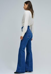 Jeans acampanados de talle alto en denim azul oscuro, con bolsillos traseros y un acabado limpio. Combinados con una blusa blanca de manga larga con detalles de botones.