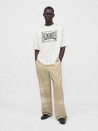Άνδρας με άσπρο oversized T-shirt με κείμενο "PALM ANGELS", μπεζ παντελόνι με φαρδιά μπότα και μαύρα παπούτσια, στέκεται με τα χέρια στις τσέπες.