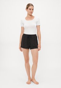 Kvinna som står barfota i en vit t-shirt med kort ärm som är åtsittande och svarta shorts med dragsko med ett subtilt leopardsmönster.