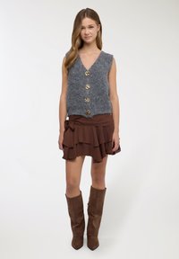 Gilet senza maniche in maglia grigia con cinque bottoni dorati, abbinato a una minigonna marrone con volant e stivali marroni in suede al ginocchio.
