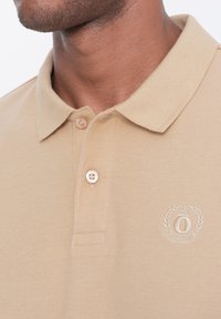 Beige polo tričko s texturovaným pleteným vzorem, klasickým límečkem, dvěma knoflíky a vyšitým logem s vavřínovými akcenty.