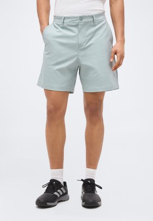 Mężczyzna w jasnoszarych szortach z bocznymi kieszeniami, białych skarpetkach i czarnych sportowych butach na jasnym, jednolitym tle.