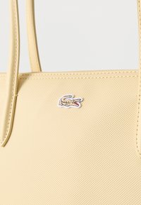 Sac cabas beige avec une surface texturée et des poignées cousues, ornées d'un petit logo crocodile brodé près du centre supérieur.