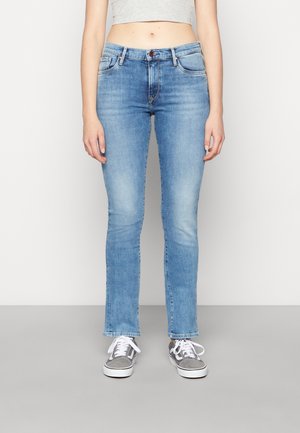 Kobieta ubrana w jasno-niebieskie obcisłe dżinsy i szaro-białe tenisówki, stojąca z ramionami luźno opartymi wzdłuż ciała na jednolitym tle.