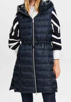 Kobieta ubrana w długi granatowy pikowany bezrękawnik z kapturem, zapinany na zamek z przodu i ze ściągaczem, nałożony na czarno-biały sweter w paski.