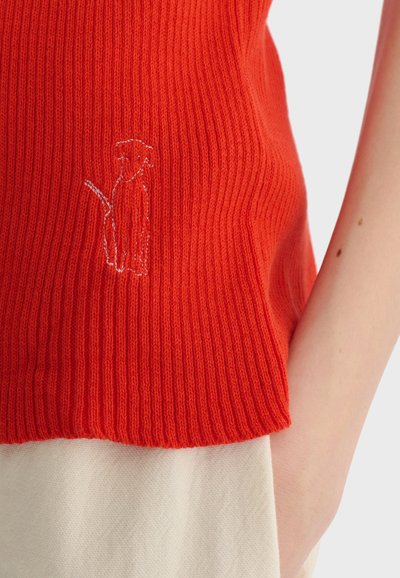 Gros plan sur un pull rouge côtelé avec le contour blanc cousu d’un chien, porté par une personne avec un bras visible et un pantalon beige.