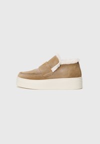 Pantofi slip-on din suede maro, cu o căptușeală pufoasă albă și o talpă din crem, având o curea Velcro unică pe partea superioară.