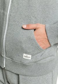 Sudadera gris con cremallera, fabricada con una mezcla de algodón. Presenta un bolsillo tipo canguro y una pequeña etiqueta con el logo de Quiksilver en el dobladillo. Textura suave.