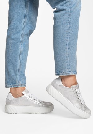 Person trägt hellblaue Jeans und metallisch silberne Sneaker mit weißen Sohlen, ein Fuß hebt sich in einem Zwischenschritt vor weißem Hintergrund.