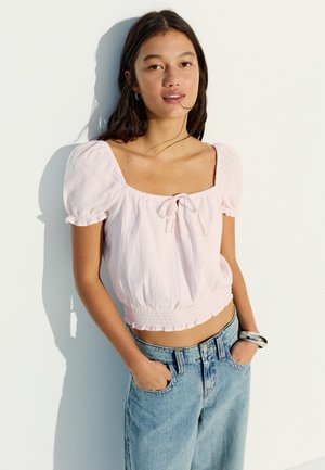 Mladá žena v světle růžovém krátkém tričku s nadýchanými rukávy a modrých džínách s vysokým pasem stojí s rukama v kapsách u bílé zdi.