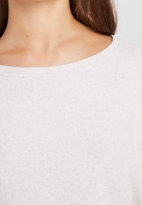 Pull tricoté beige clair avec un large décolleté et une texture douce, présentant un design simple sans motifs ni éléments visibles.