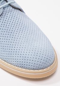 Chaussure en daim bleu clair avec texture perforée, bout arrondi, semelle en caoutchouc beige et couture blanche autour du bord.