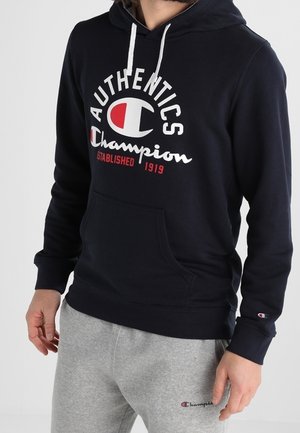 Mann, der eine marineblaue Champion-Kapuze mit weißen Kordeln und graue Champion-Sweatpants trägt, beide mit Markenlogos versehen.