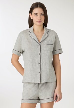 Giovane donna che indossa un pigiama a maniche corte color grigio chiaro con bottoni e pantaloncini abbinati con rifiniture scure, in piedi davanti a uno sfondo bianco.