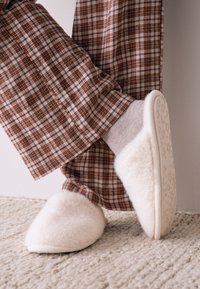 Chaussons doux de couleur crème avec une texture pelucheuse et un bout arrondi, associés à un pantalon à carreaux marron et crème. Des chaussettes tricotées sont visibles.