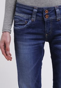Denim jeans in dark blue with subtle fading, featuring a classic button closure, two front pockets, and a fitted silhouette.