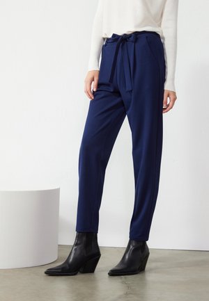 Femme portant un pantalon bleu marine à la taille nouée, un haut blanc à manches longues et des bottines à talons noires, se tenant à l'intérieur sur un sol en béton.