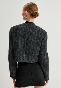 Zwarte cropped jacket van textuurstof met verticale patronen. Het heeft lange mouwen en een kraagloos ontwerp, gecombineerd met een zwarte rok.