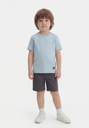 Jonge jongen staand, draagt een lichtblauw t-shirt met geel logo, donkergrijze korte broek, witte sokken en witte sneakers tegen een effen witte achtergrond.