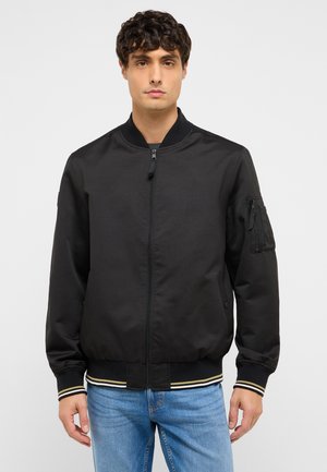 Junger Mann trägt eine schwarze Bomberjacke mit Reißverschluss und gestreiften Bündchen, kombiniert mit hellblauen Jeans, stehend vor einem schlichten Hintergrund.