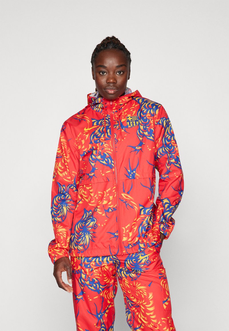 Chaqueta roja con un patrón floral azul y amarillo, con cremallera en el frente, capucha y puños elásticos. Combinada con pantalones a juego, todos con un diseño similar.