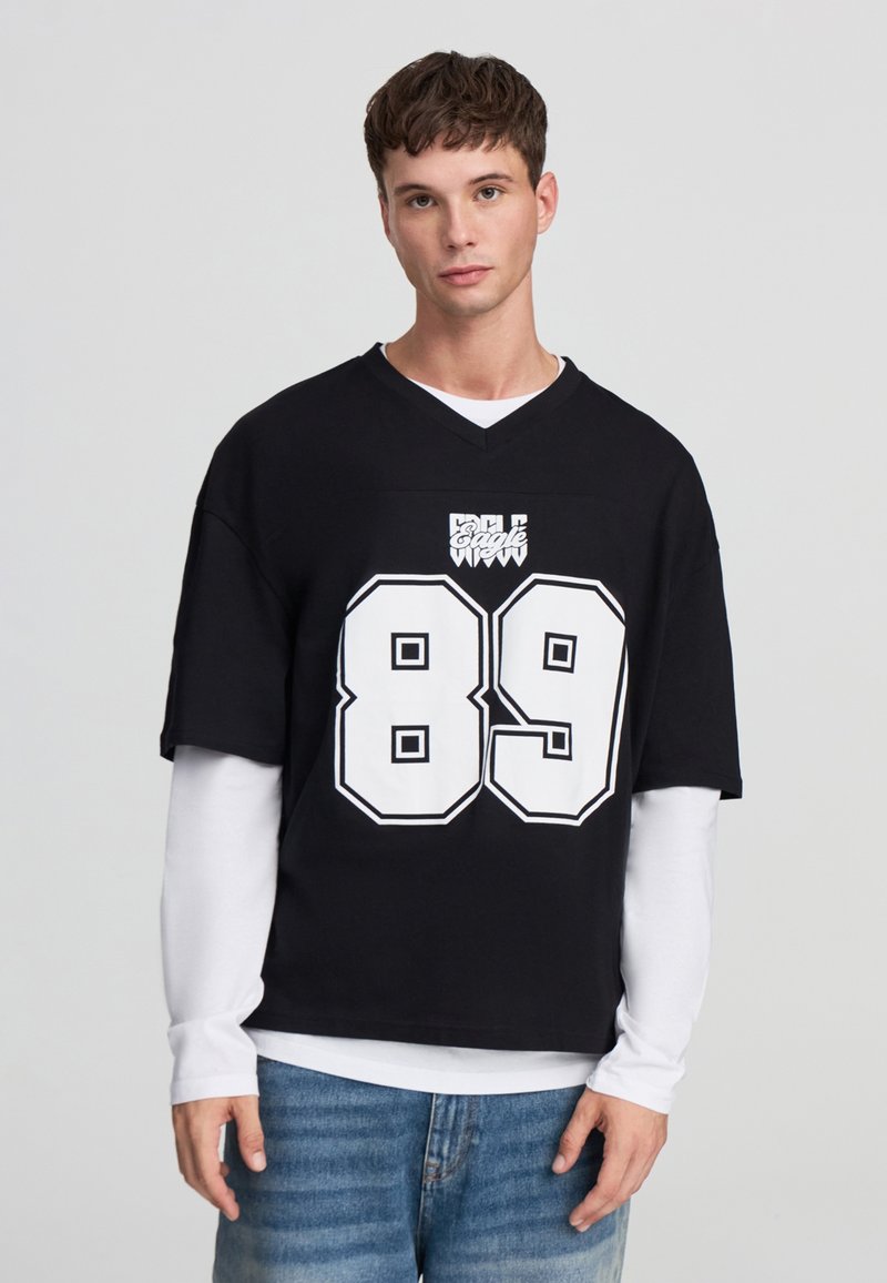 Jersey nera oversize a maniche corte con grafica e logo bianchi "89". Indossato sopra una maglietta bianca a maniche lunghe, abbinato a jeans azzurri chiari.