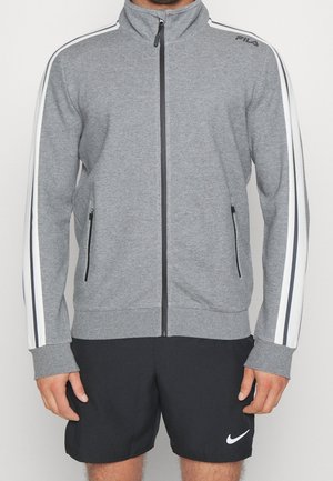 Homme portant une veste grise zippée avec des rayures blanches et noires sur les manches et un short noir avec le logo Nike blanc.
