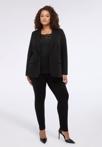 Blazer nero con due tasche frontali, abbinato a un top nero con bordo in pizzo e pantaloni neri aderenti, completato da scarpe nere con tacco alto.
