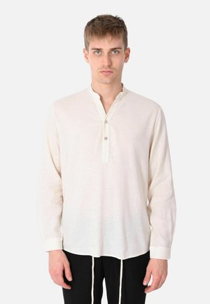 Giovane uomo che indossa una camicia beige larga a maniche lunghe con colletto abbottonato, in piedi davanti a uno sfondo chiaro uniforme.