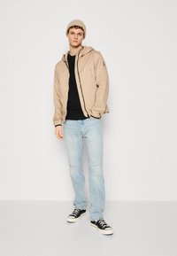 Beige hooded jacket with a zipper and black accents, paired with a black shirt and light blue distressed jeans, finished with black sneakers.
