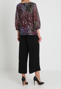 Blouse fleurie transparente avec motifs violets, noirs et blancs, associée à un pantalon large noir et des escarpins noirs à talons aiguilles, avec une coupe ample.