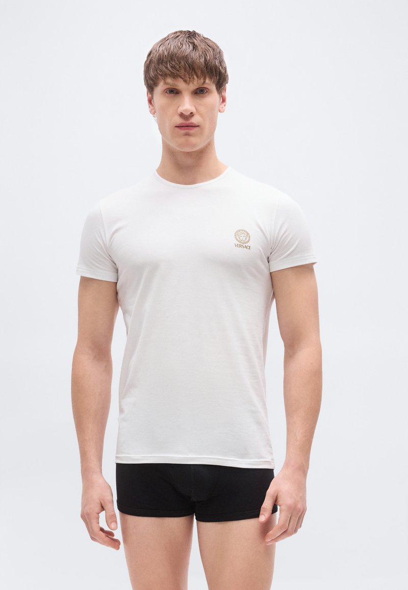Λευκό κοντομάνικο T-shirt με στρογγυλ ή λαιμόκοψη και χρυσό λογότυπο Versace στο αριστερό στήθος, σε συνδυασμό με μαύρο κοντό σορτς. Λείο ύφασμα.