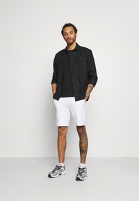 Camisa negra de botones sobre una camiseta negra, pantalones cortos blancos y zapatillas en blanco y negro. Tatuajes destacados en una pierna.