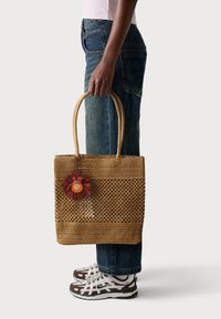 Geflochtene Strohtasche mit einem runden floralen Akzent in Rot und Orange. Verfügt über robuste Obergriffe und ein offenes Design für bessere Sichtbarkeit.