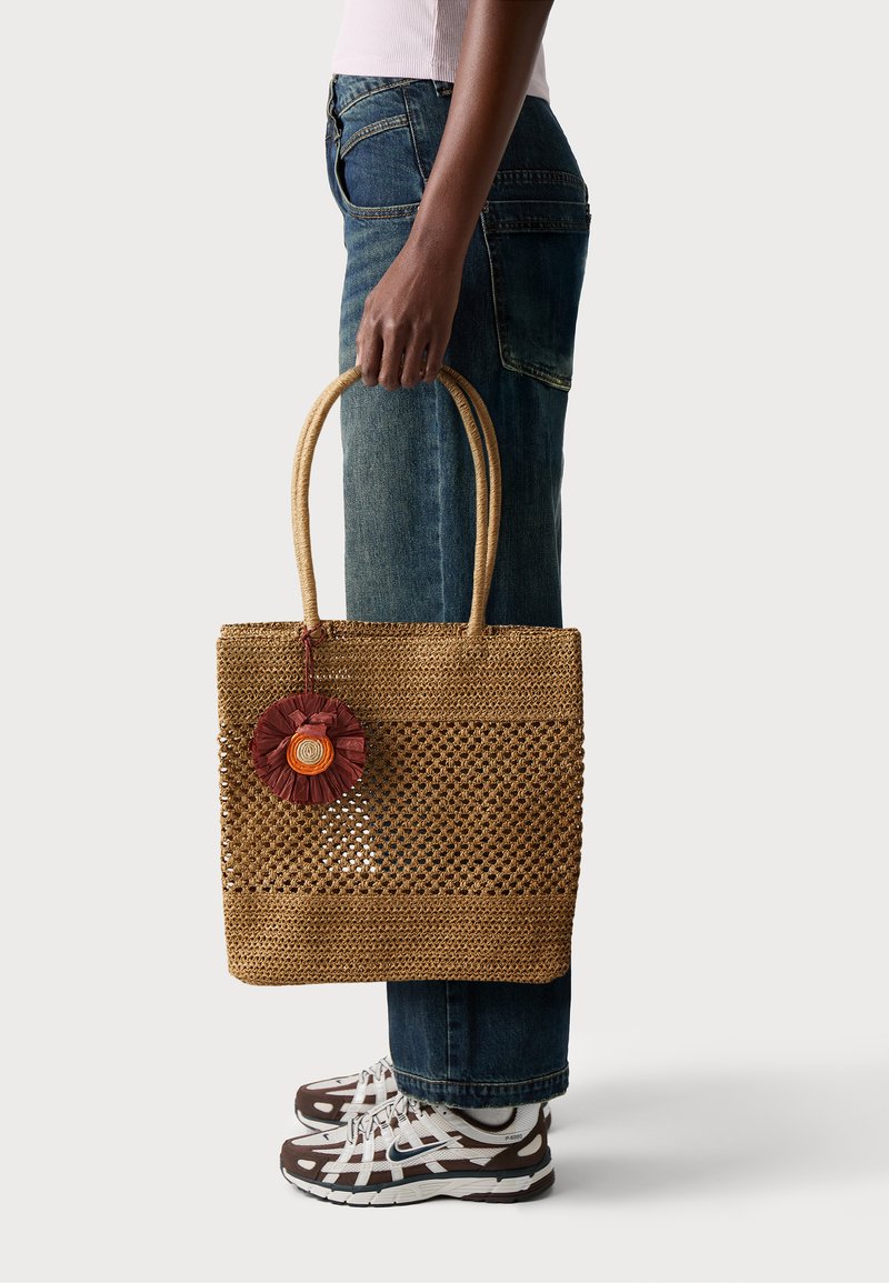Geflochtene Strohtasche mit einem runden floralen Akzent in Rot und Orange. Verfügt über robuste Obergriffe und ein offenes Design für bessere Sichtbarkeit.