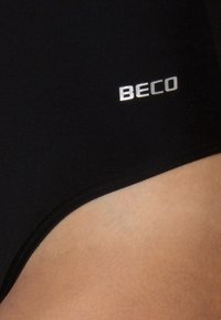 Maillot de bain noir avec une texture lisse, présentant un logo "BECO" en argent sur le côté. Le design est simple, sans motifs ni accents supplémentaires.