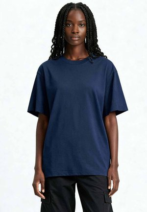 Donna con lunghi capelli intrecciati che indossa una T-shirt oversize blu scuro semplice e pantaloni cargo neri, in piedi contro uno sfondo bianco.