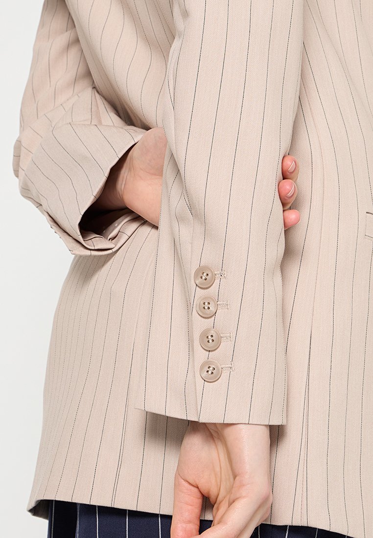Manteau beige à rayures avec des manches ornées de quatre boutons beiges, une main glissée sous l'autre bras derrière le dos.