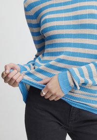 Blå och beige randig stickad tröja med ribbad textur, åtsittande ärmar och en något lös passform, tillsammans med mörka byxor.