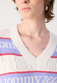 V-hals trui in roomkleur, lavendel en rood. Heeft een kabelbreipatroon, logo-accenten en een geribde halslijn. Textuurdetail van de stof is zichtbaar.