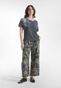 Ung kvinna som står med händerna i fickorna, iklädd en mörk t-shirt med blommönster, gröna vida blommiga byxor och bruna snörskor.