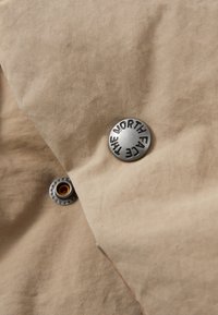 Beige fabric with a smooth texture, featuring a round silver button with "THE NORTH FACE" engraved, and a smaller metal snap detail.