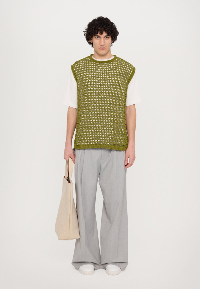 Grünes, gestricktes, ärmelloses Oberteil mit lockerer Passform, kombiniert mit grauen, weiten Hosen und weißen Sneakers; hält eine beigen Tragetasche.