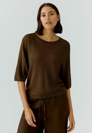 Frau mit glatten dunklen Haaren, die ein braunes Strickoberteil mit Ellbogenärmeln und passende Hosen mit Kordelzug vor hellem Hintergrund trägt.