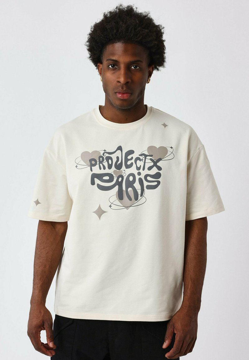 Camiseta de gran tamaño en color crema, fabricada con un tejido suave, que presenta un gran gráfico gris con acentos de corazones y estrellas. Manga corta, cuello redondo.