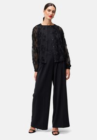 Schwarze, bestickte transparente Jacke über einem passenden Oberteil und weitem Hosenbein. Glatter Stoff mit floralen Details, kombiniert mit schwarzen Sandalen.